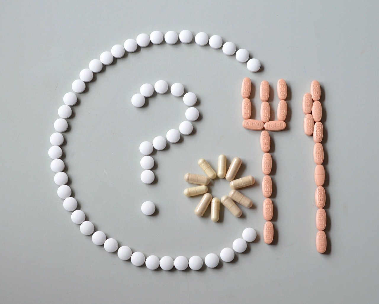 Arrangement of various pills and capsules forming a question mark, fork, and sun shape, symbolizing